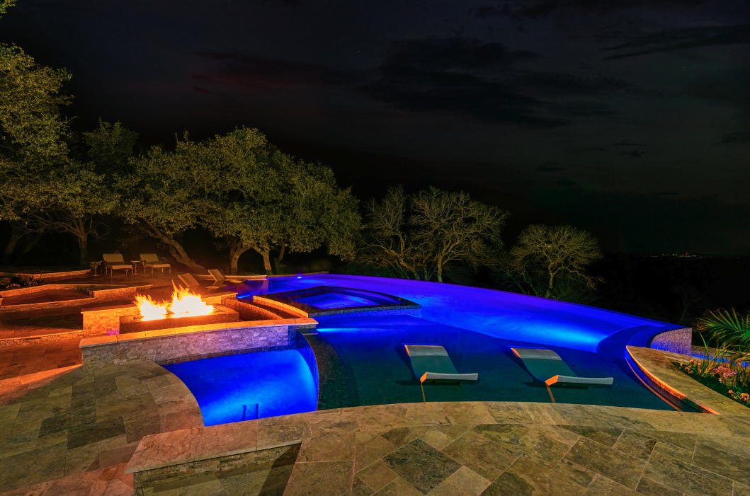 Nighttime view of a luxury infinity pool in the Texas Hill Country featuring vibrant blue LED lighting, a stone fire feature, in-pool loungers, and natural travertine decking designed by Infinity Pools of Texas